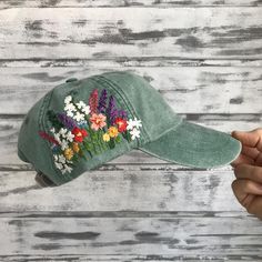 a green hat with colorful flowers embroidered on the front and side, held up against a white wooden background