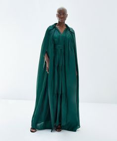 This luxurious sleeveless cape is crafted from soft satin fabric. It boasts a gathered neckline, open front, and sparkling rhinestone buttons that add a touch of elegance. Pair it with the Andile dress (sold separately) for a stunning evening ensemble. One size fits most. Meet the Designer: The Baruni collection offers beautiful silhouettes and sophisticated fabrics as unique as you, for every occasion. Fadwa Baruni’s use of unique texture and color in her flowing fabrics allow her designs to st Luxury Designer Dresses With Front Open, Luxury Front Open Designer Dresses, Luxury Green Thobe For Wedding, Luxury Evening Dress With Gathered Neckline, Luxury Open Front Evening Dresses, Luxury Green Kaftan With Traditional Drape, Luxury Designer Front Open Dresses, Luxury Gathered Neckline Evening Dress, Silk Cape Dress For Gala