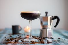 A coupe cocktail glass filled with an espresso martini, blurry items on the background: a mocha pot, a small burning candle and an espresso cup. They all sit on a blue wooden table with coffee beans coffee particles spread on the table. Winter Mocktails, Martini Espresso, Espresso Martini Cocktail, Cinnamon Simple Syrup, White Cranberry Juice, Jack Rose, Fall Cocktails, Coffee Uses