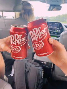 two people holding up dr pepper soda cans in the back seat of a car,