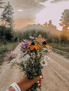 a person holding flowers in their hand on the side of a dirt road at sunset