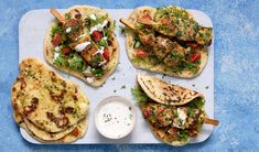 four pita breads with different toppings on a blue tablecloth next to a small bowl of ranch dressing