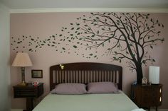 a bed sitting under a tree painted on the wall next to a nightstand and lamp