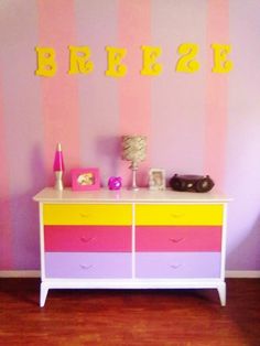 a room with pink, yellow and purple stripes on the wall next to a dresser