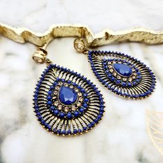 Captivating in its royal essence, this jewelry set features a striking combination of deep blue stones and gold-toned metalwork. The necklace, with its simple yet elegant golden bead chain, leads to a pendant boasting a rich blue stone at its core, encircled by a halo of darker blue crystals. The showstoppers are the chandelier earrings, presenting an intricate peacock feather-inspired design with a teardrop blue stone centerpiece, framed by layers of fine, dark blue crystals that create a drama Elegant Blue Stone Earrings, Elegant Blue Earrings With Stones, Elegant Blue Filigree Necklace, Blue Beaded Brass Necklaces, Sapphire Metal Jewelry For Party, Gold Teardrop Pendant Jewelry For Party, Party Brass Jewelry With Gold Beads, Teardrop Pendant Metal Jewelry For Parties, Antique Gold Bohemian Jewelry For Party