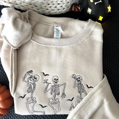 Forget everything you think you know about spooky season style. This sweatshirt isn't just cozy, it's got the best moves! Picture this: You're cuddling up with a pumpkin spice latte, but with a twist - this embroidered skeleton crew is breakin' it down on the front, reminding you that even the afterlife has rhythm. ☁️ Amazingly Soft and Comfort to wear  🧵 Stitched in my home, each sweatshirts may vary ever so slightly. 🛍️ Product Details: Sizes Available: Unisex sizing from a range of sizes to Halloween Sweatshirt Diy, Cute Halloween Sweaters, Halloween Embroidered Sweatshirt, Embroidered Skeleton, Fall Crewneck Sweatshirt, Embroidered Halloween, Sorority Sweatshirts, Fall Apparel, Dancing Skeletons