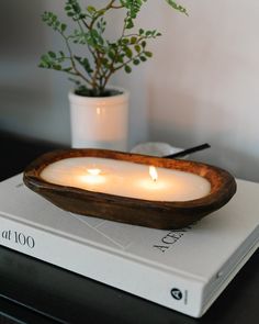 two lit candles in a wooden bowl on top of a white book next to a potted plant