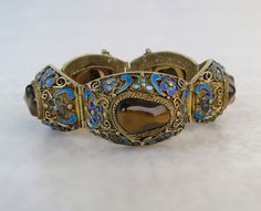 Smooth chatoyant tigers eye gemstones are centered on the ornate enameled links of this antique Chinese gilt silver bracelet. This bracelet was likely a Chinese export piece circa 1920s, although similar examples were made as far back as the 1890s. The large 1 1/2 inch links are constructed with intricate mesh filigree metal work. The links are decorated with cannetille flowers topped with colorful enamel and golden curls. There is a gold vermeil finish to the silver giving the bracelet an overa Traditional Brown Bangle Jewelry, Antique Jeweled Bangle Jewelry, Antique Jeweled Bangle, Ornate Cabochon Bracelet Jewelry, Ornate Cabochon Bracelet, Antique Enamel Bracelet Jewelry, Tigers Eye Bracelet, Tigers Eye Gem, Tigers Eye Stone