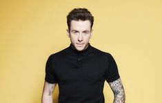 a man with tattoos standing in front of a yellow wall wearing a black polo shirt