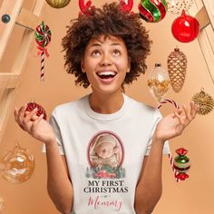 a woman is holding up christmas ornaments and looking at the camera while wearing a t - shirt that says, my first christmas mommy