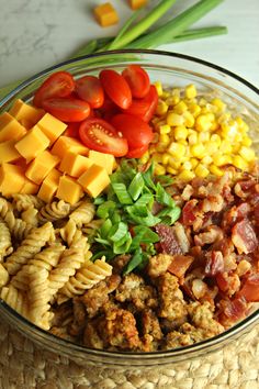 a glass bowl filled with pasta, tomatoes, corn and other vegetables on top of a table