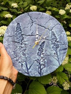 a hand holding up a blue clock in front of some bushes and flowers with white flowers around it