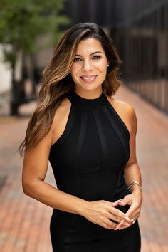 a beautiful woman in a black dress posing for a photo on a brick sidewalk with trees and buildings behind her