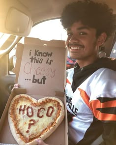 a man holding up a heart shaped pizza in the back seat of a car with an i know this is cheesy but hoco written on it