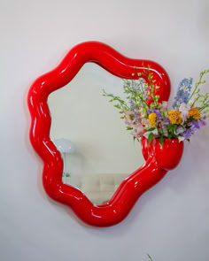 a red vase with flowers in it sitting next to a mirror on a white wall