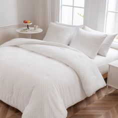 a bed with white comforter and pillows in a room next to a window on a wooden floor