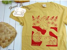 "= Rabbit Hole = They have a playroom, a powder room, and a nursery Rabbit Hole \" 100% American Grow Cotton Unisex T-shirt. This soft garment-dyed bunny tee, illustrated and hand-printed with Silk Screen by Mina Wilcox, was created with pure joy from her small art studio in California. We only use non-toxic water-based inks, ensuring they are safe for you and the Earth. Made from 100% ring-spun cotton, responsibly sourced from American farms, and carefully garment-dyed, this tee exudes a retro look and feel. DETAILS: One 100% American Grow Cotton Garment Dyed Short Sleeve T-shir *Garment dyeing offers a remarkable method for creating trendy and captivating apparel with a beloved, well-worn t-shirt appearance. This aesthetic is achieved by applying the dye to the clothing after it has unde Grow Cotton, Small Art Studio, Relax Lifestyle, Bunny T Shirt, Toxic Water, Tshirt Art, Pure Joy, Hand Screen Printed, Rabbit Hole