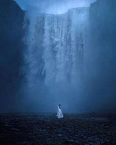 a woman standing in front of a waterfall under a cloudy sky with her back to the camera