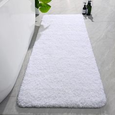 a white bathroom rug on the floor next to a bathtub and toilet in front of a plant