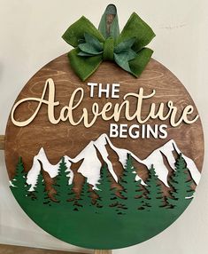 a wooden sign that says the adventure begins with mountains and evergreens on it in front of a white wall