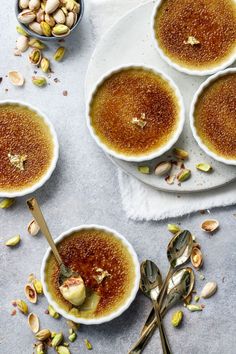 four white bowls filled with food on top of a table next to spoons and nuts