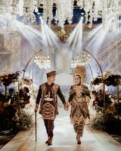 two people walking down a runway with chandeliers hanging from the ceiling behind them