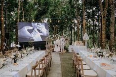 an outdoor wedding setup with white linens and greenery on the tables, surrounded by tall trees