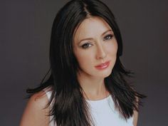 a woman with long black hair wearing a white top and posing for a photo in front of a gray background