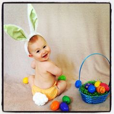 a baby wearing an easter bunny costume next to a basket full of eggs and colorful candies