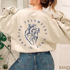 This Cardiac Telemetry Unit Sweatshirt / Telemetry Nurse Sweater is perfect for work! Cozy and warm, it makes a great gift too!   **FRONT & BACK PRINT**  **UNISEX SIZING** ♥ W E L C O M E  T O  S K E T C H Y  C A T  D E S I G N S ! ♥ Click here to return to our shop's home page ⇒ https://www.etsy.com/shop/SketchyCatDesigns Say hello to your new favorite sweatshirt! All products are printed with eco-friendly water-based inks, giving them the softest feel. The design itself is embedded into the fa Cardiac Nurse Sweatshirt, Med Surg Nurse Shirt, Nurse Sweatshirt Ideas, Nursing Art, Telemetry Nursing, Cardiac Nurse, Nurse Sweater, Cardiac Nursing, Nurse Shirts