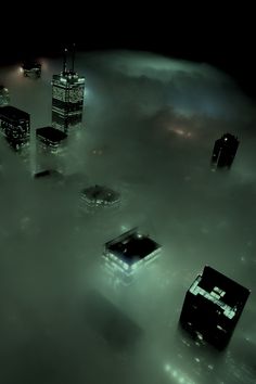 an aerial view of skyscrapers in the foggy sky at night with lights shining on them