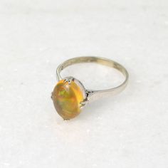 "A rainbow is swimming in this gorgeous antique opal ring! A beautifully proportioned opal cabochon is centered with cardinal prongs (north, south, west and east) in an 18k white gold setting. The opal features a transparent orange body, typical of Mexican jelly opals, with a \"contra luz\" color flash seen when the gem interacts with light. The color play is astounding, with every color of the rainbow ignited within the body of the opal, having painterly brush-stroke like patterns. This ring hails from the 1920s and has been tenderly well-cared for in its lifetime. It is in wonderful condition and ready for a new owner. This ring could be a perfect alternative engagement ring, gift for someone with an October birthstone or simply a present for an antique jewelry lover.   Opal is a fascina Oval Cabochon Opal Ring With Polished Finish, Fine Jewelry Oval Cabochon Opal Ring, Ethiopian Opal Oval Cabochon Ring In Yellow Gold, Yellow Gold Ethiopian Opal Oval Cabochon Ring, Oval Cabochon Opal Ring Fine Jewelry, Classic Oval Opal Ring Collectible, Vintage Oval Moonstone Ring With Polished Finish, Oval Cabochon Opal Ring In Fine Jewelry, Yellow Gold Ethiopian Opal Ring In Oval Cabochon Shape