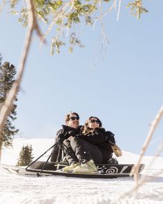 two people sitting in the snow with skis on their feet and one person wearing sunglasses