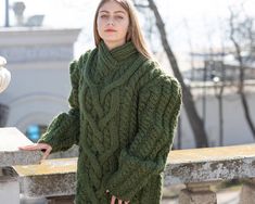 a woman wearing a green cabled sweater standing next to a stone wall and looking off into the distance