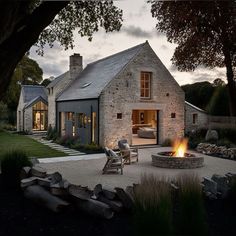 a stone house with a fire pit in the front yard at dusk, surrounded by grass and trees