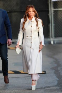 a woman in a white coat and heels walks down the street with a man behind her