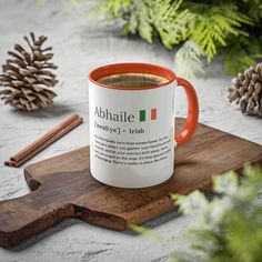 an orange and white coffee mug sitting on top of a wooden cutting board next to pine cones