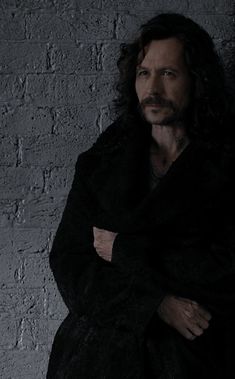 a man with long hair standing in front of a brick wall wearing a black coat