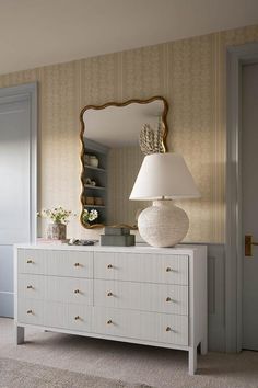 a white dresser sitting next to a lamp and a mirror on top of a wall