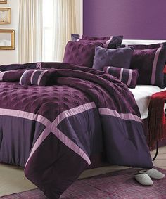 a bed with purple comforter and pillows in a room next to a lamp on a table