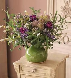 a green vase filled with lots of flowers on top of a wooden table next to a dresser