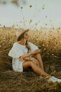 a woman sitting in the middle of a field wearing a white dress and hat with her legs crossed