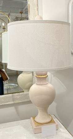 a white lamp sitting on top of a table next to a mirror with a woman in the background