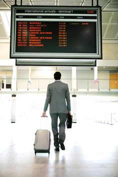 a man in a suit and tie pulling a suitcase through an airport with information on the wall behind him