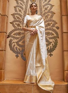 a woman wearing a white and gold sari standing in front of a decorative wall