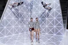 two models walk down the runway in front of an array of white lights and geometric shapes