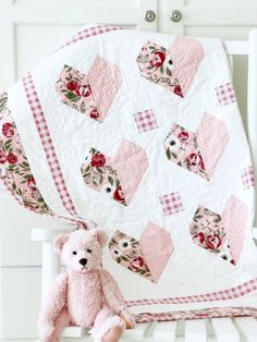 a pink teddy bear sitting on top of a white chair next to a quilted blanket