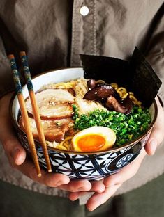 a person holding a bowl of food with chopsticks and an egg on top