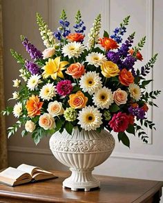 a white vase filled with lots of colorful flowers on top of a table next to an open book