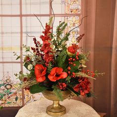 a gold vase with red flowers and greenery on a table in front of a window